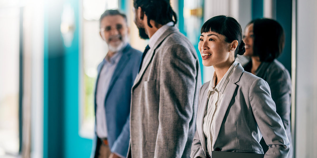 A group of business professionals dressed in formal attire standing in a brightly lit modern office space. The setting features teal walls and large windows, creating a professional yet vibrant atmosphere. The individuals are engaged in conversation or ob